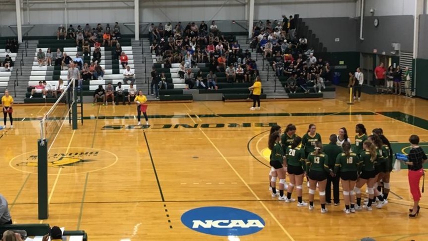 The volleyball team huddles during a timeout in front of a large crowd.
